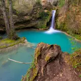 Cascada de agua turquesa en la ruta del agua Berganzo, rodeada de vegetación y rocas cubiertas de musgo.