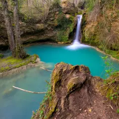 Cascada de agua turquesa en la ruta del agua Berganzo, rodeada de vegetación y rocas cubiertas de musgo.