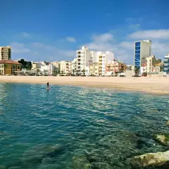Un día soleado en la costa, con un paddleboarder solitario en el mar turquesa y edificios modernos de fondo. ¡Un oasis de playas cerca de Zaragoza!