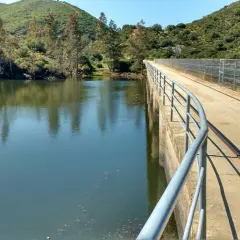 Un puente de hormigón con barandilla recorre un embalse, ideal para una ruta senderismo Sevilla. El agua refleja árboles y colinas verdes.
