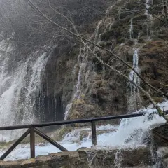 Cascada en la ruta nacimiento río Cuervo. Agua cayendo sobre rocas cubiertas de musgo y vegetación. Barandilla de madera en primer plano.