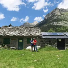 Pareja posa frente a una rústica cabaña de turmo con paneles solares, rodeada de exuberante vegetación y majestuosas montañas.