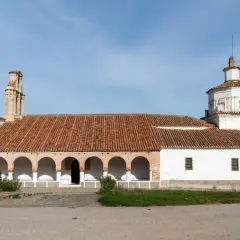 La ermita Virgen del Ara, con su tejado de tejas rojas y arcos blancos, se alza bajo un cielo azul.