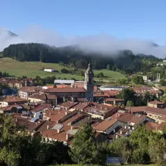 Vista panorámica de Oñati, con su iglesia y tejados rojizos, rodeada de verdes colinas y niebla. Ideal para planificar cómo llegar.