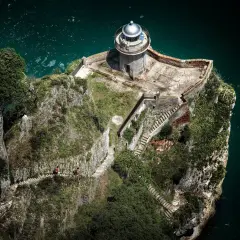 El faro del caballo se alza sobre un acantilado rocoso. Dos personas caminan por un sendero empinado hacia él.