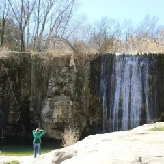 Una persona admira una cascada en un paisaje rocoso. El agua cae sobre la pared de roca, creando un velo. El sol brilla, y la vegetación seca rodea la escena. Pozan de Vero, un lugar de belleza natural.