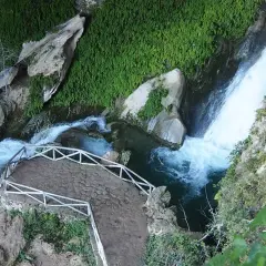 Sendero de piedra y barandilla blanca junto a la cascada de la cueva del agua Tiscar, rodeada de vegetación exuberante.