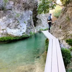 Senderista cruza un puente de madera sobre el agua cristalina en el Parrizal de Beceite, rodeado de rocas y vegetación exuberante.
