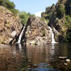 Dos cascadas caen por rocas escarpadas en la cascada del hervidero, formando un estanque oscuro. Personas observan desde lo alto.