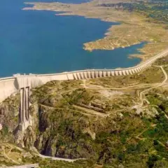 Vista aérea de la imponente presa de Almendra, reteniendo un vasto embalse de agua azul. La estructura curva se alza majestuosa sobre un paisaje rocoso y verde.