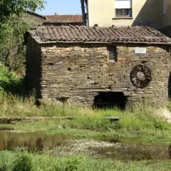 Antiguo molino de agua de piedra, con su rueda oxidada y vegetación exuberante a su alrededor.