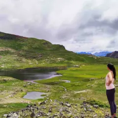 Mujer contempla los ibones de Anayet, lagos de montaña rodeados de verdes prados y picos rocosos bajo un cielo nublado.