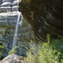 Una fina cascada de la mea cae por una pared rocosa, rodeada de vegetación exuberante y rocas cubiertas de musgo.