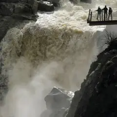 Tres personas observan la furia del pozo de los humos desde una pasarela. El agua turbia cae con fuerza, creando una densa niebla.