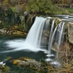 La majestuosa cascada de Tobalina, con su velo de agua cayendo sobre rocas cubiertas de musgo, rodeada de árboles otoñales y un río sereno.