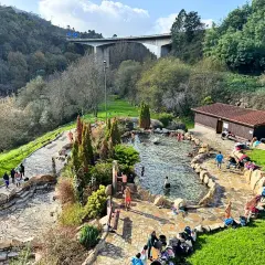 Gente disfrutando de las **termas en Galicia**, un oasis de relajación rodeado de naturaleza, con un puente moderno al fondo.