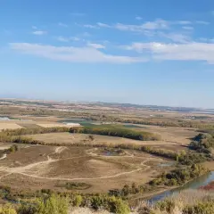 Paisaje rural con un río serpenteante cerca de Villafranca del Bierzo. Campos cultivados y árboles de otoño bajo un cielo azul con nubes.