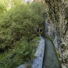 Senderistas caminan por la ruta de los bolos, un sendero estrecho junto a un canal de agua y un acantilado rocoso.