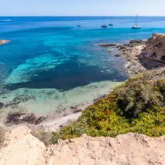 Aguas turquesas y arena blanca en una cala rocosa, un paraíso entre las mejores playas de Formentera. Yates navegan en el horizonte.