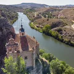 Ermita de San Saturio, Soria, se alza sobre un acantilado junto al río Duero, rodeada de vegetación y colinas.