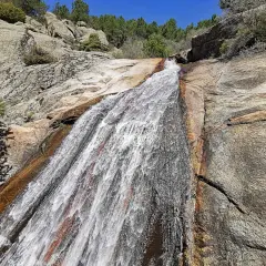 El agua desciende por la roca en la cascada del Hornillo, rodeada de vegetación y rocas graníticas bajo un cielo azul.