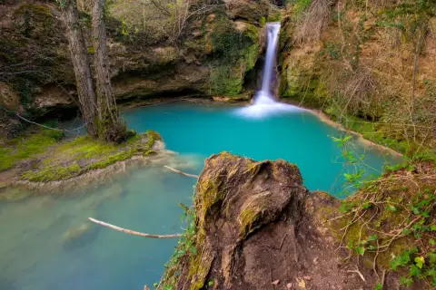 Cascada de agua turquesa en la ruta del agua Berganzo, rodeada de vegetación y rocas cubiertas de musgo.