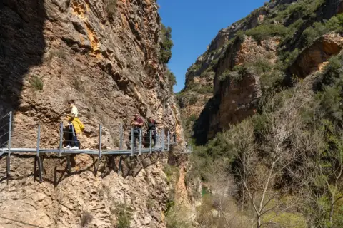 Gente camina por las pasarelas de Alquézar, un sendero suspendido entre imponentes paredes rocosas y vegetación.