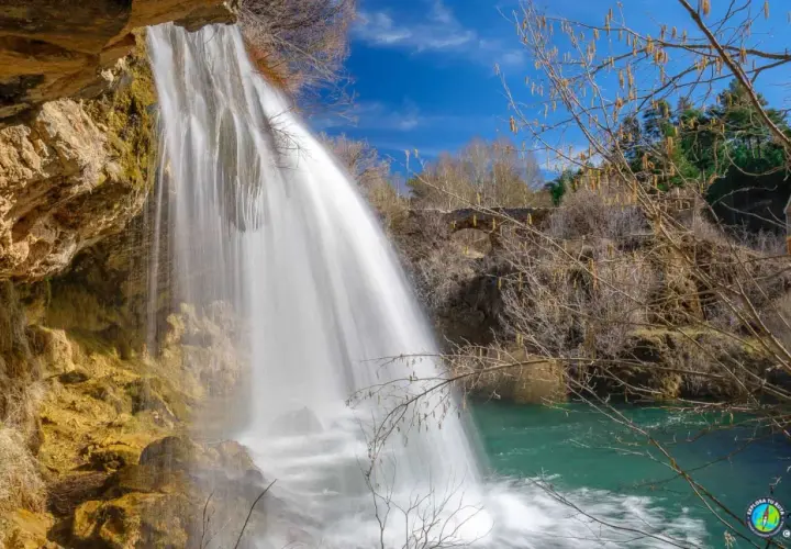 Cascada de agua blanca cayendo sobre rocas ocres, como los ojos del Cabriel, en un día azul.