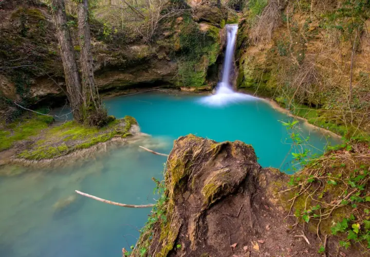 Cascada de agua turquesa en la ruta del agua Berganzo, rodeada de vegetación y rocas cubiertas de musgo.