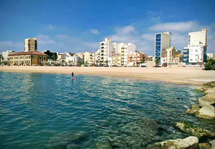 Un día soleado en la costa, con un paddleboarder solitario en el mar turquesa y edificios modernos de fondo. ¡Un oasis de playas cerca de Zaragoza!