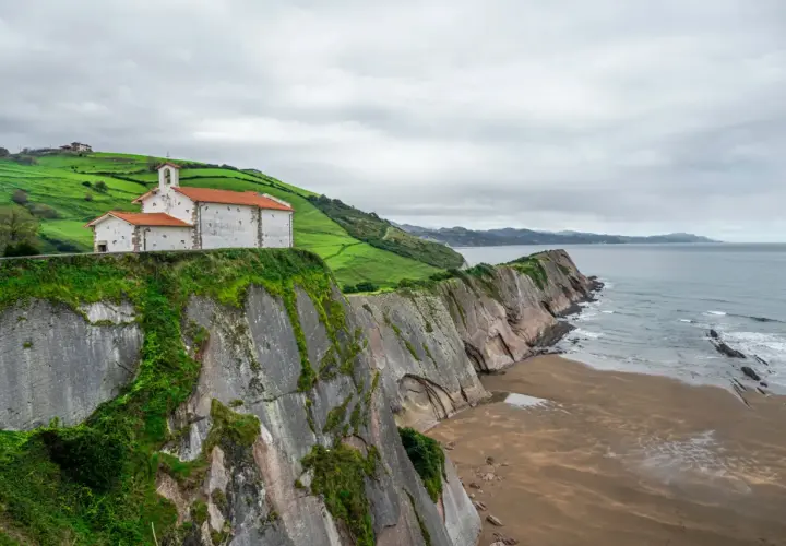 Iglesia blanca con tejado naranja sobre acantilado verde, junto al mar Cantábrico. Un lugar que evoca la esencia de "8 apellidos vascos".