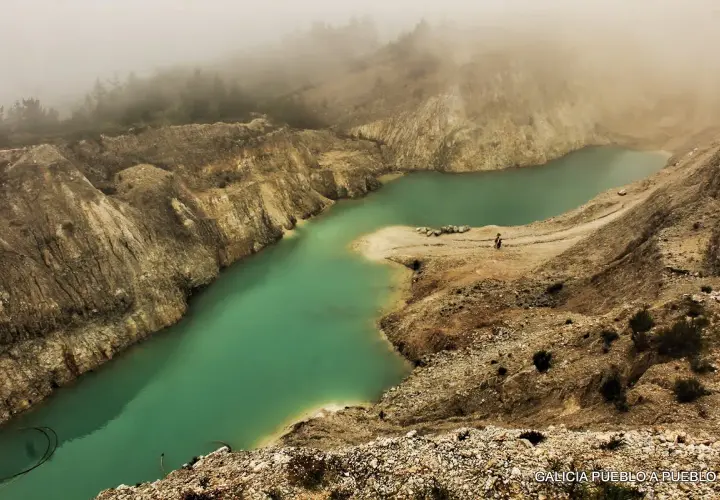 Laguna turquesa en el monte Neme Carballo, rodeada de tierra y rocas, con niebla al fondo.