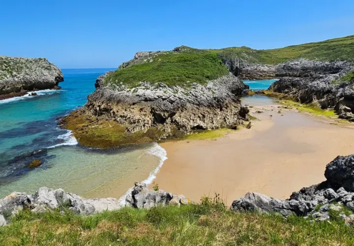 Una cala escondida entre acantilados verdes, con aguas turquesas y arena dorada. Un rincón idílico entre las mejores playas de Asturias.