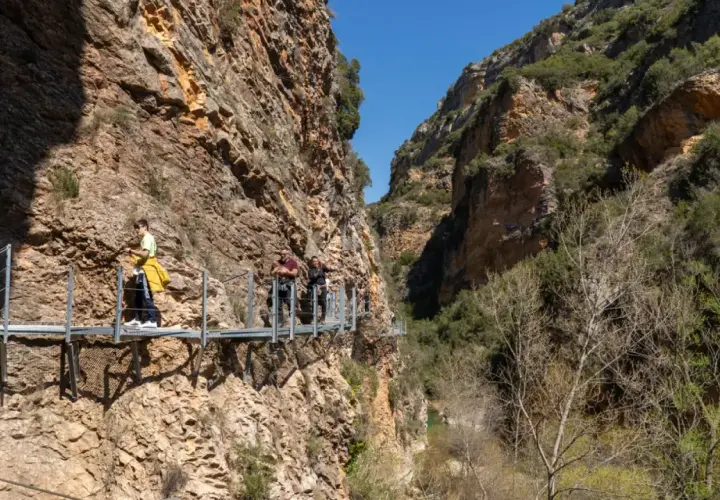 Gente camina por las pasarelas de Alquézar, un sendero suspendido entre imponentes paredes rocosas y vegetación.