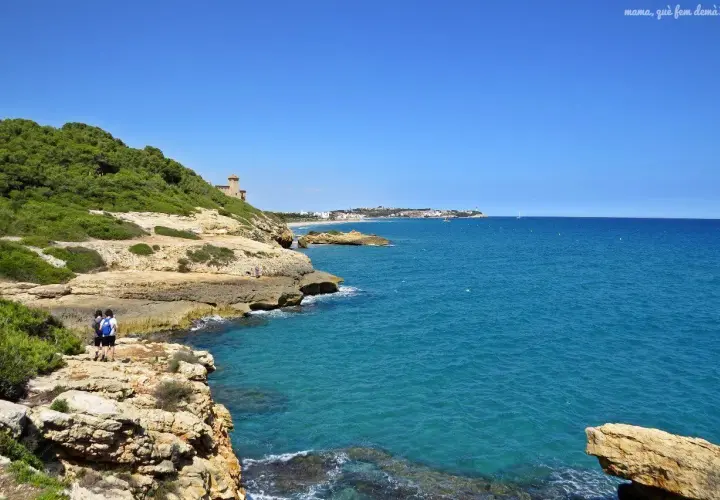 Dos personas caminan por el camí de ronda de Tarragona, disfrutando de las vistas al mar azul y la costa rocosa.