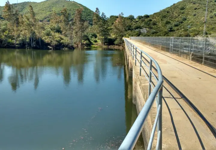 Un puente de hormigón con barandilla recorre un embalse, ideal para una ruta senderismo Sevilla. El agua refleja árboles y colinas verdes.