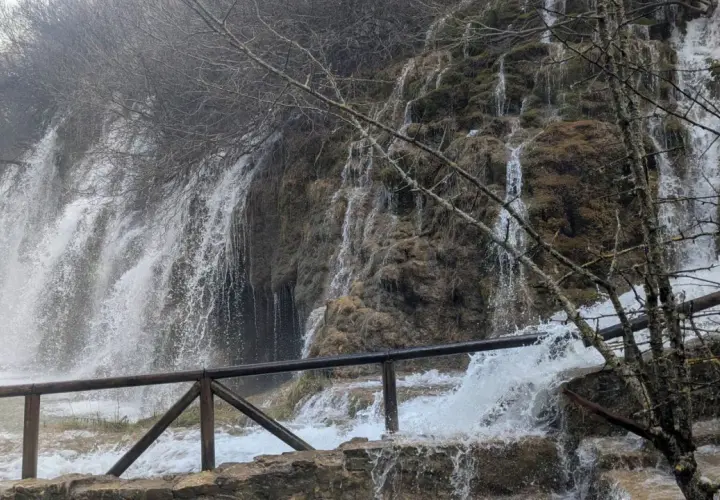 Cascada en la ruta nacimiento río Cuervo. Agua cayendo sobre rocas cubiertas de musgo y vegetación. Barandilla de madera en primer plano.