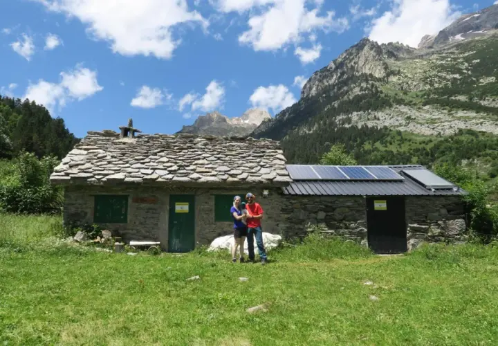 Pareja posa frente a una rústica cabaña de turmo con paneles solares, rodeada de exuberante vegetación y majestuosas montañas.