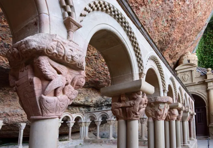 Detalles arquitectónicos y escultóricos en piedra de San Juan de la Peña, con arcos y capiteles tallados bajo una roca.