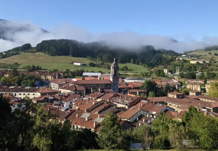 Vista panorámica de Oñati, con su iglesia y tejados rojizos, rodeada de verdes colinas y niebla. Ideal para planificar cómo llegar.