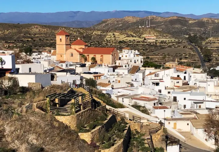 Vista panorámica de Lucainena de las Torres, un pueblo blanco andaluz con una iglesia de campanario naranja. El camino para llegar serpentea entre colinas áridas.