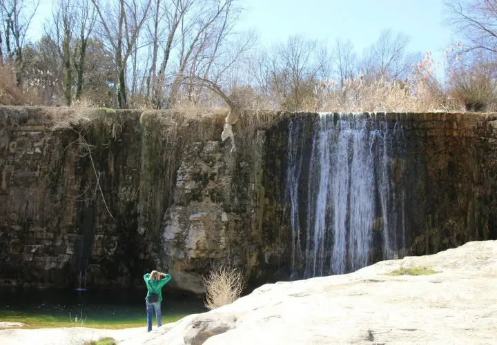 Una persona admira una cascada en un paisaje rocoso. El agua cae sobre la pared de roca, creando un velo. El sol brilla, y la vegetación seca rodea la escena. Pozan de Vero, un lugar de belleza natural.