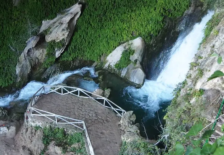 Sendero de piedra y barandilla blanca junto a la cascada de la cueva del agua Tiscar, rodeada de vegetación exuberante.