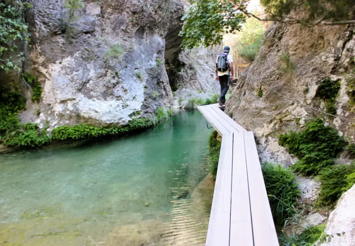 Senderista cruza un puente de madera sobre el agua cristalina en el Parrizal de Beceite, rodeado de rocas y vegetación exuberante.