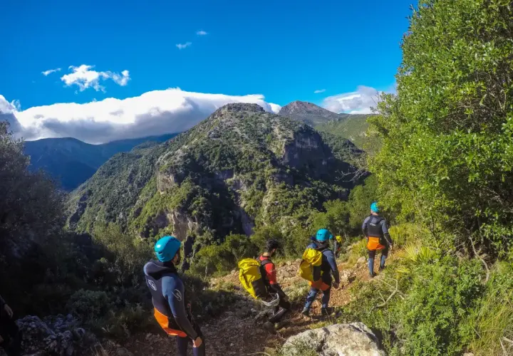 Grupo de barranquistas desciende por un sendero rocoso hacia la garganta verde, rodeados de exuberante vegetación y montañas.