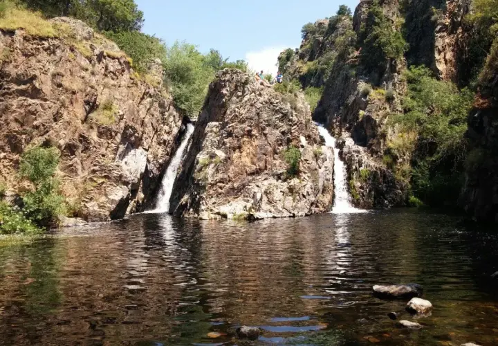 Dos cascadas caen por rocas escarpadas en la cascada del hervidero, formando un estanque oscuro. Personas observan desde lo alto.
