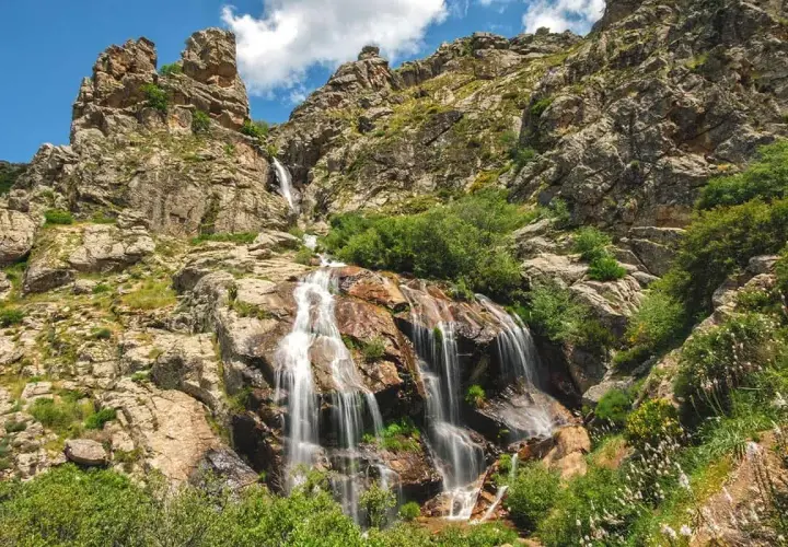 La cascada de los litueros desciende por rocas escarpadas, rodeada de vegetación exuberante bajo un cielo azul con nubes.