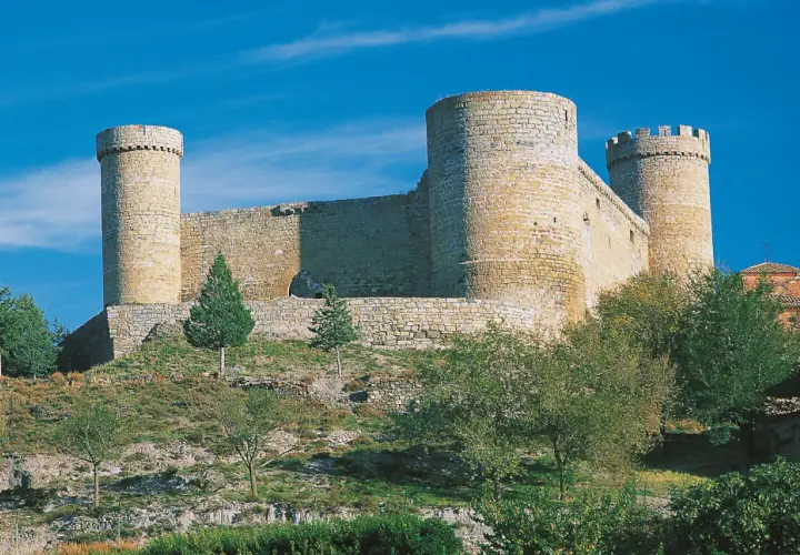 El castillo de Cornago se alza imponente sobre una colina, con sus torres circulares y murallas de piedra bajo un cielo azul intenso.