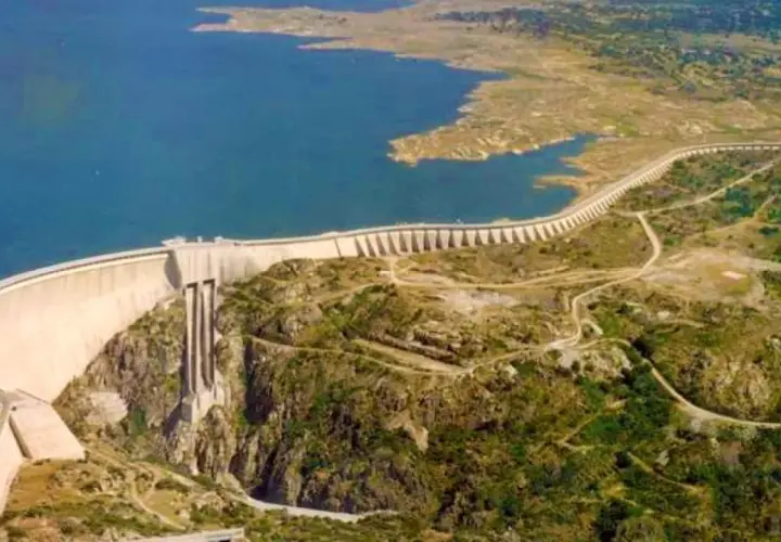 Vista aérea de la imponente presa de Almendra, reteniendo un vasto embalse de agua azul. La estructura curva se alza majestuosa sobre un paisaje rocoso y verde.