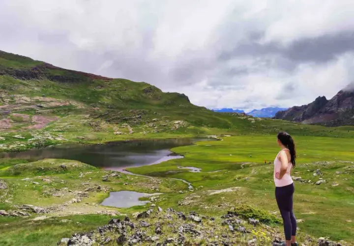 Mujer contempla los ibones de Anayet, lagos de montaña rodeados de verdes prados y picos rocosos bajo un cielo nublado.
